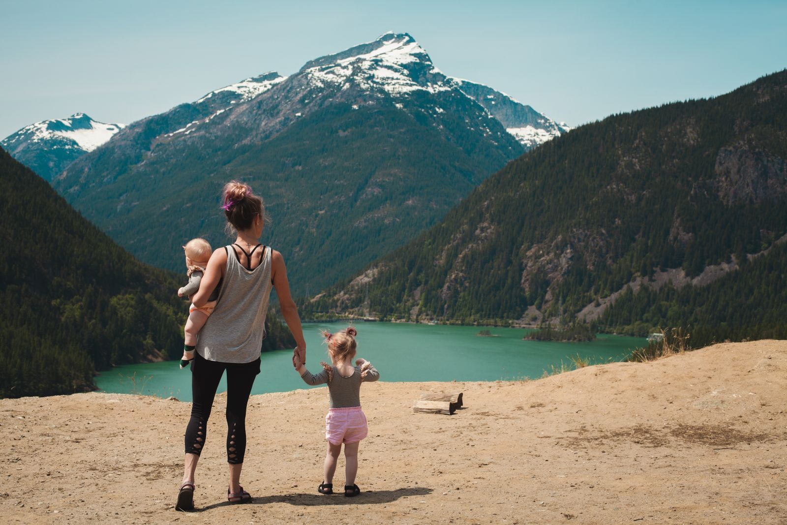 A mother and two small children standing by a turquoise mountain lake under snowy peaks
